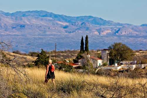 A person in a red backpack walks through tall grass with mountains and a small house in the background.