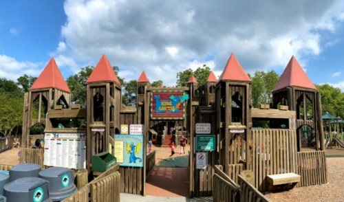 A wooden playground with colorful towers and a sign, surrounded by trees and children playing under a cloudy sky.