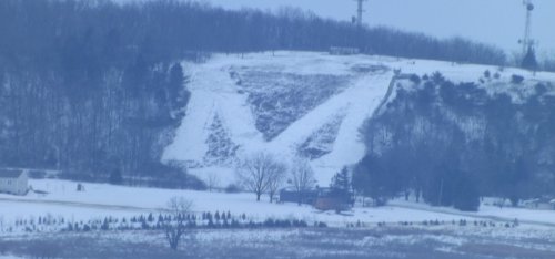 A snowy hillside features a large, carved "M" shape, surrounded by trees and a few buildings in the foreground.