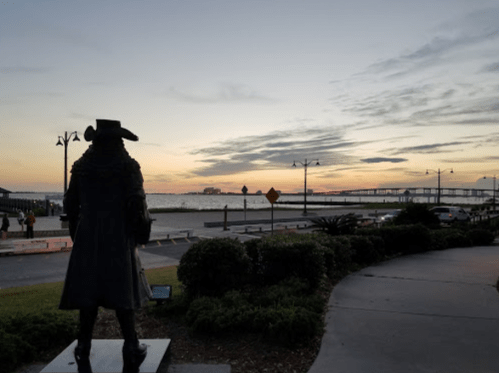 A statue of a historical figure stands by the waterfront at sunset, with a cloudy sky and distant buildings visible.