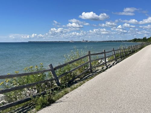 A scenic view of a calm lake with a wooden fence along a paved path, under a blue sky with fluffy clouds.