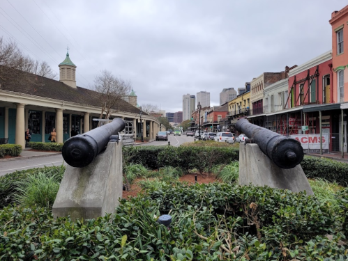 Two cannons point down a street lined with historic buildings and modern skyscrapers under a cloudy sky.