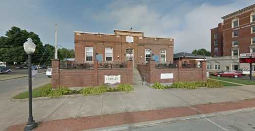 A brick library building with a sign, surrounded by greenery and a sidewalk, under a cloudy sky.