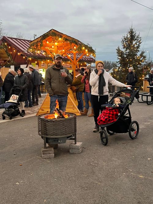 A couple stands by a fire pit, holding drinks, while a child in a stroller is wrapped in a blanket at a festive outdoor market.