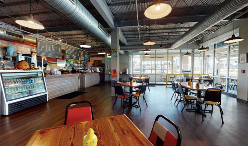 A spacious restaurant interior with wooden tables, chairs, and a counter displaying food and drinks. Bright and inviting atmosphere.