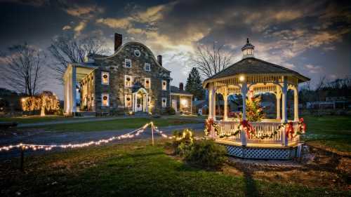 A historic stone house and gazebo decorated for the holidays, with festive lights and a Christmas tree at dusk.