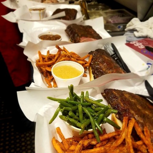 A variety of barbecue dishes with ribs, green beans, sweet potato fries, and dipping sauce on white paper plates.
