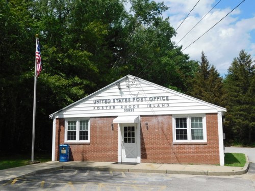 A small brick building with a sign reading "United States Post Office" and an American flag outside.