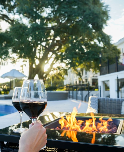 Two glasses of red wine are held up near a fire pit, with a pool and trees in the background during sunset.