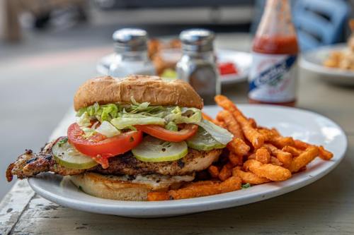 A grilled chicken sandwich with lettuce, tomato, and pickles, served with sweet potato fries and condiments on the side.
