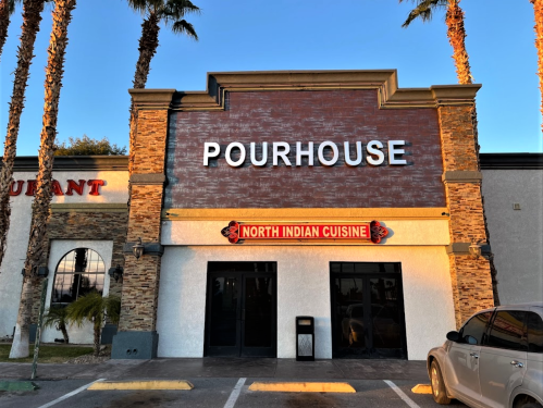 Exterior of a restaurant building with "POURHOUSE" sign and "NORTH INDIAN CUISINE" banner, surrounded by palm trees.