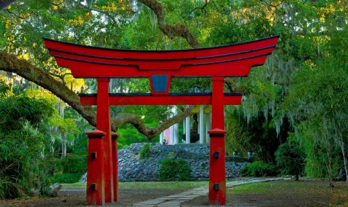 A vibrant red torii gate stands in a lush green garden, framed by trees and moss, inviting visitors to explore.