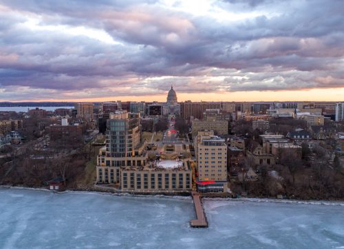 Aerial view of a city skyline at sunset, with buildings near a frozen lake and dramatic clouds overhead.