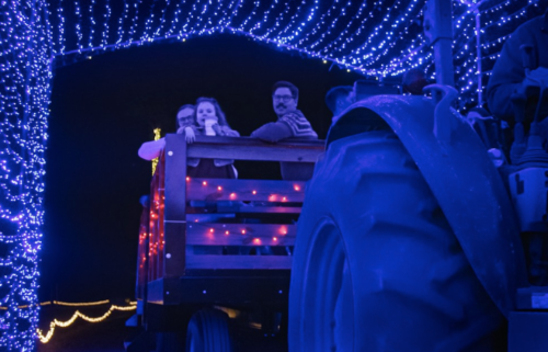 A tractor pulls a festive wagon decorated with colorful lights, with people enjoying the ride.
