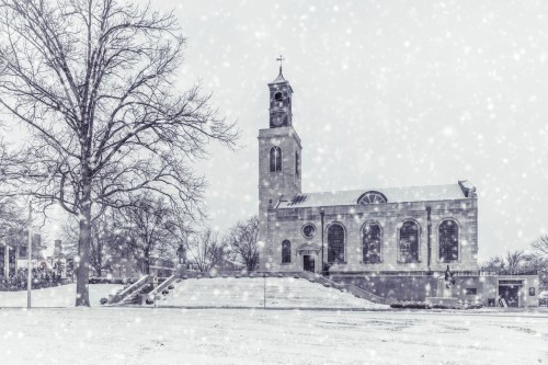 A snow-covered church stands amidst falling snowflakes, surrounded by bare trees and a quiet winter landscape.