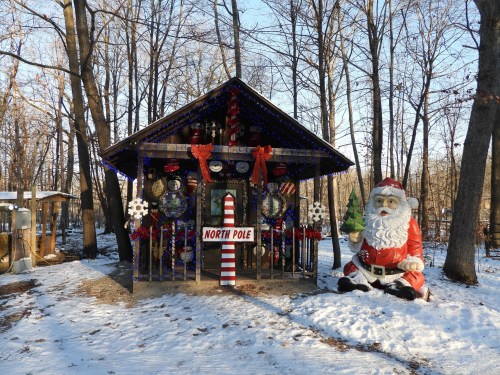 A festive cabin decorated for Christmas, featuring a large Santa statue and a "North Pole" sign, surrounded by snow and trees.
