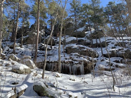 A snowy landscape with rocky cliffs and tall trees under a clear blue sky. Icicles hang from the rocks.