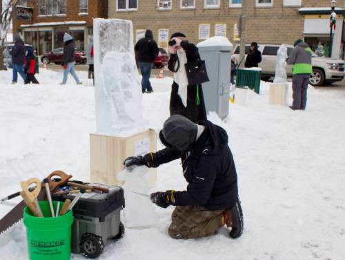 A person chisels an ice sculpture while another observes, surrounded by snow and tools in a winter festival setting.