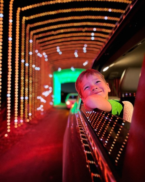 A smiling child leans out of a car window, surrounded by colorful lights in a festive tunnel.