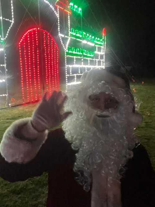 A person dressed as Santa Claus waves in front of a brightly lit festive display at night.