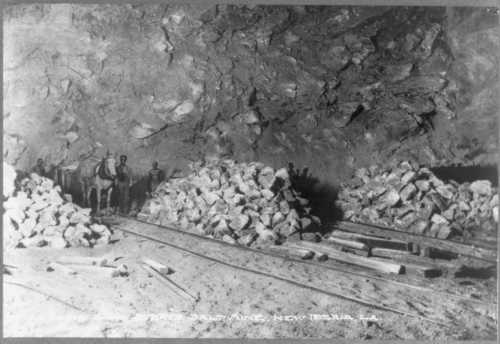 Black and white photo of miners and a horse in a mine, with stacked rocks and wooden beams in the foreground.