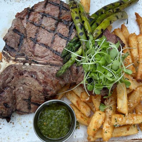 A grilled steak with asparagus, microgreens, and crispy fries, served with a side of green sauce on a white plate.