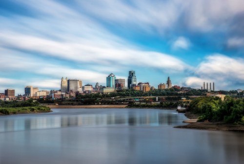 A scenic view of a city skyline reflected in a calm river under a blue sky with wispy clouds.