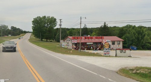 A roadside store with a red roof, signs advertising food, and trees in the background along a two-lane highway.