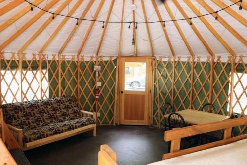 Interior of a yurt featuring a wooden frame, a sofa, a dining table, and string lights.