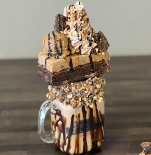 A tall milkshake topped with whipped cream, brownies, and chocolate drizzle, served in a glass mug.