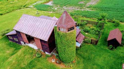 Aerial view of a rustic barn with a tower covered in greenery, surrounded by lush fields and a garden.