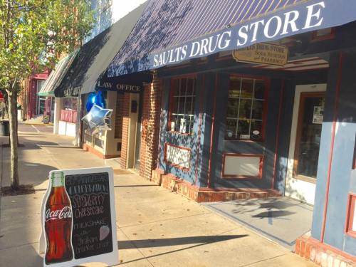 A storefront with a blue awning, featuring a sign for Saults Drug Store and a chalkboard advertising strawberry cheesecake.