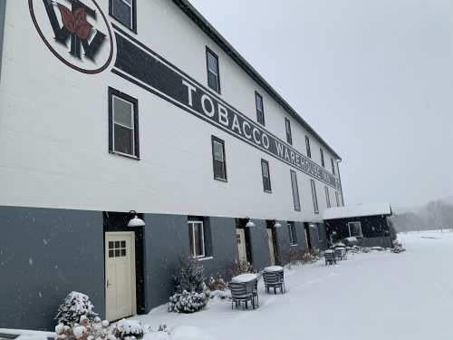 A snowy scene of a tobacco warehouse building with a large sign, surrounded by a white landscape and outdoor seating.