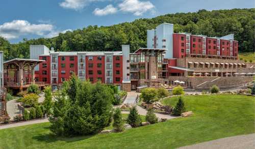 A modern hotel building with red and stone exterior, surrounded by greenery and hills under a blue sky.