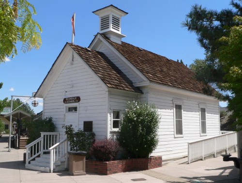 A small white historic building with a peaked roof, surrounded by greenery and a clear blue sky.