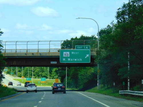 Highway sign indicating Exit 1A for Route 113 West toward West Warwick, with cars driving below.