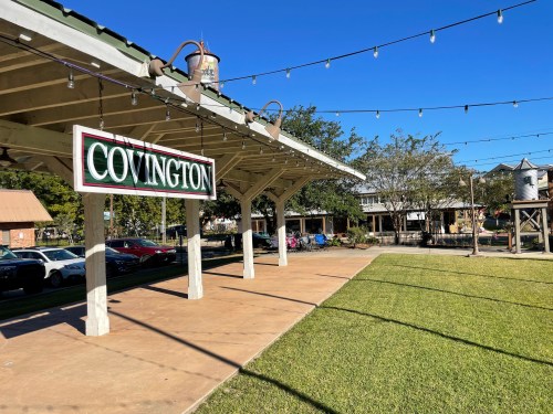 A covered pavilion with a "COVINGTON" sign, string lights, and a grassy area in a town square setting.