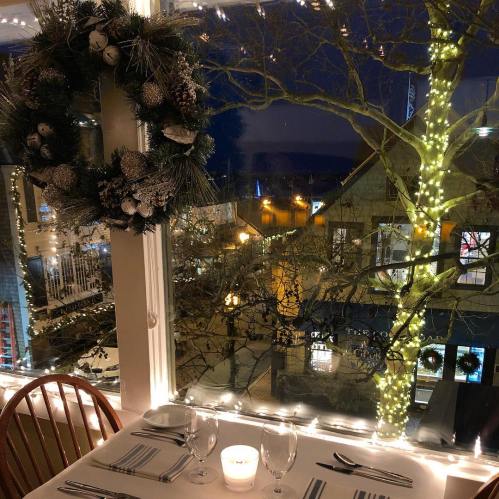 A cozy restaurant table by a window, decorated with a festive wreath and soft candlelight, overlooking a twinkling street.