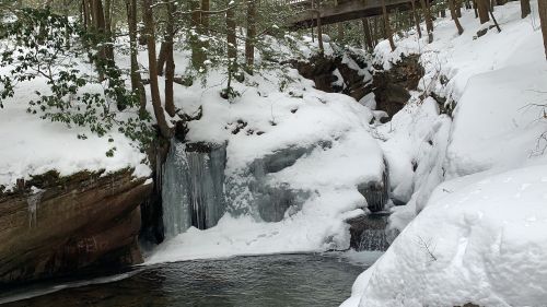 A snowy landscape featuring a frozen waterfall and a stream, surrounded by trees and a wooden bridge above.