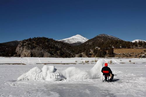 A person in a red jacket sits on a chair by a snow sculpture on a frozen lake, surrounded by mountains under a clear blue sky.