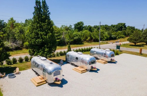 Three shiny Airstream trailers parked on a gravel lot, surrounded by greenery and a clear blue sky.