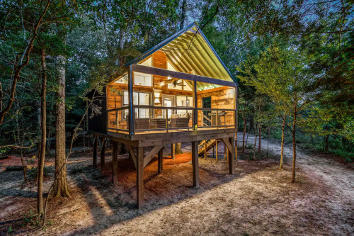 A modern cabin on stilts surrounded by trees, illuminated at dusk with large windows and a welcoming porch.