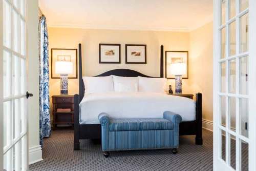 A cozy hotel room featuring a large bed, two lamps, and a striped bench at the foot, with framed artwork on the walls.