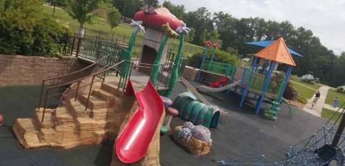 Colorful playground featuring slides, climbing structures, and a playhouse surrounded by green trees and open space.