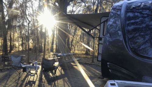 A camper parked under trees with sunlight streaming through, featuring outdoor chairs and a table set up nearby.