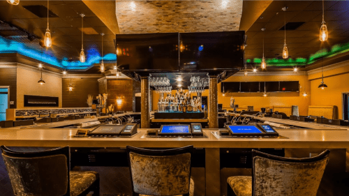 Modern bar interior with sleek seating, illuminated shelves, and a large screen above the counter.