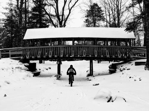 A cyclist rides under a snow-covered wooden bridge in a winter landscape. Trees surround the scene.