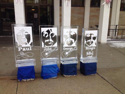 Four ice sculptures featuring faces and names of Paul, John, George, and Ringo, displayed outside a building.