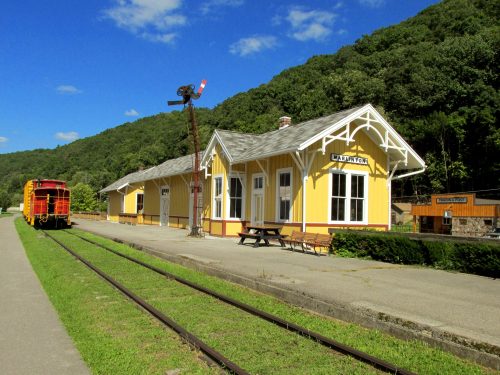 A yellow train station building with a red train nearby, set against a green hillside under a blue sky.