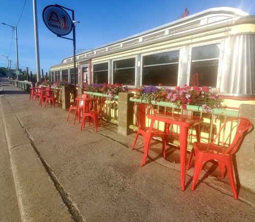 A colorful diner with red chairs and tables outside, adorned with flowers, under a clear blue sky.